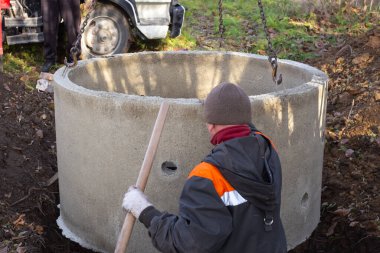 Doldurucu, foseptik tankı yapmak için bir çukura beton halkayı indiriyor. Bir işçi yere kanalizasyon kuruyor..
