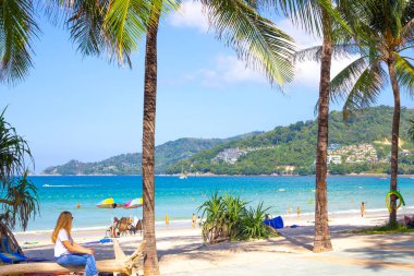 Phuket, Tayland-12.29.2018: Güneşli bir günde deniz ve dağa bakan palmiyeli sahil Patong Plajı, turistler dinleniyor.