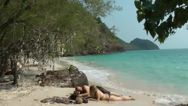 Un hombre y una mujer rubia en traje de baño yacen en la arena blanca, en la orilla del mar con olas en una laguna aislada. Viajes y turismo en los trópicos. — Vídeo de stock