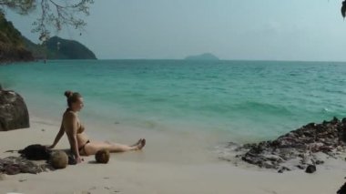 Sarı saçlı Avrupalı bir kadın, mayo giymiş bir topuz içinde gelen bir dalgayla kumlu bir deniz kıyısında oturuyor, tropik bir gölde, görünüşü ve gülüşü var..