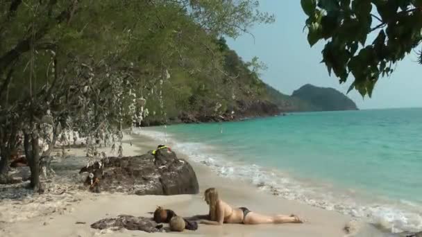 Une Européenne blonde aux cheveux longs en maillot de bain se trouve sur le sable au bord de la mer, baignée par une vague transparente. Concept de voyage et de tourisme. — Video