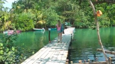 Uzun saçlı sarışın bir kadın, tropiklerdeki bir nehrin üzerindeki ahşap köprü boyunca yürür, yeşil bankalarla birlikte. Seyahat ve turizm.