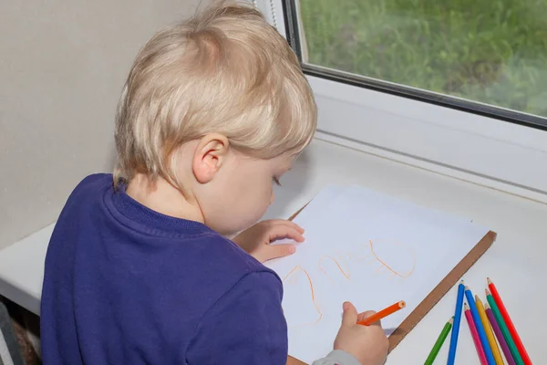 Boy blond draws with a pencil on a white sheet of paper while sitting by the window. Fun for children.