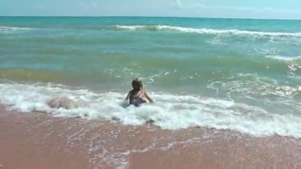 Uma menina de fato de banho brinca numa praia de areia com uma onda que se aproxima. Férias de verão no mar. — Vídeo de Stock