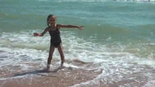 Ein kleines Mädchen im Badeanzug spielt an einem Sandstrand mit einer entgegenkommenden Welle. Sommerurlaub am Meer. — Stockvideo