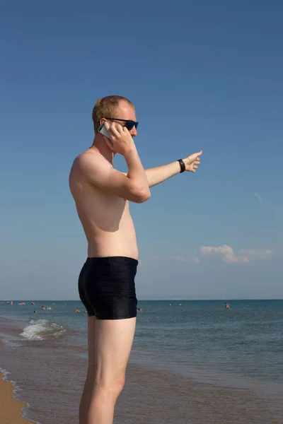 裸の胴を持つ青年が海岸に立って電話で話しています — ストック写真