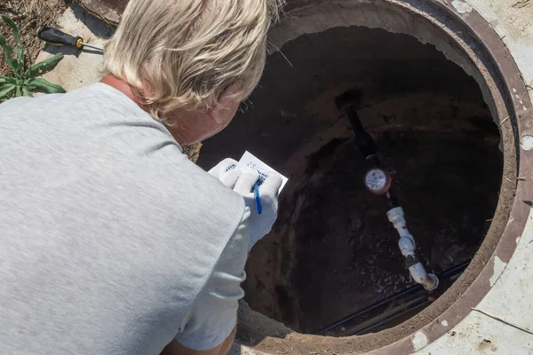 Mannen som böjer sig över vattenbrunnen och registrerar vattenmätarens värden. Kontroll och fixering av räknaren — Stockfoto