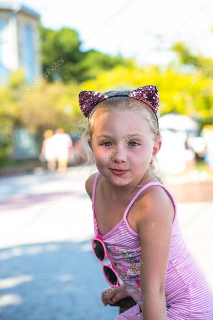 A little blonde girl with cat ears on her head looks at the camera and smiles. Summer fun holidays