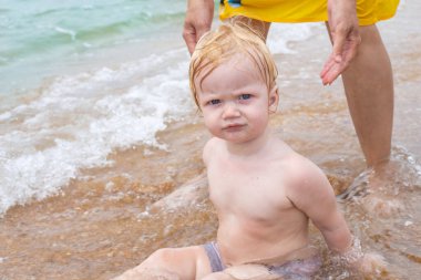 Küçük sarışın bir çocuk deniz dalgası ile kumda annesinin yanında oturuyor. Suda yaz tatili.
