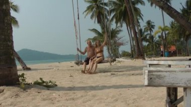 Bir adam ve mayo giymiş bir kadın deniz kıyısındaki palmiye ağacına asılı salıncakta sallanıyorlar. Tropikal sahilde dinlenme ve dinlenme