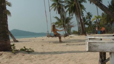 Uzun saçlı, mayo giymiş bir sarışın palmiye ağacından sarkan bir salıncakta sallanıyor. Güzel tropikal deniz manzarası