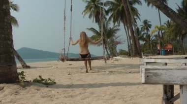 Uzun saçlı, mayo giymiş bir sarışın palmiye ağacından sarkan salıncakta oturur ve palmiye ağaçlarına ve denize bakarak sallanır.