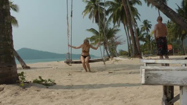 Una mujer en traje de baño se levanta de un columpio suspendido en una palmera y se acerca a un hombre en la orilla del mar. Descanso y relajación en la costa tropical — Vídeos de Stock