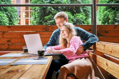 Laptopun arkasındaki kafede mutlu genç bir çift. Kız ve erkek gülümseyerek eğleniyorlar.
