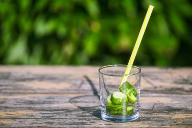 Yazın ferahlatıcı bir içki hazırlıyorum. Dilimlenmiş salatalıkla dilimlenmiş su olmayan cam, yeşil doğanın arka planına karşı ahşap bir masada.