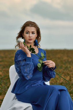 Romantik kadın portresi. Mavi elbiseli kadın tarlada sandalyede otururken elinde beyaz bir gül tutuyor.