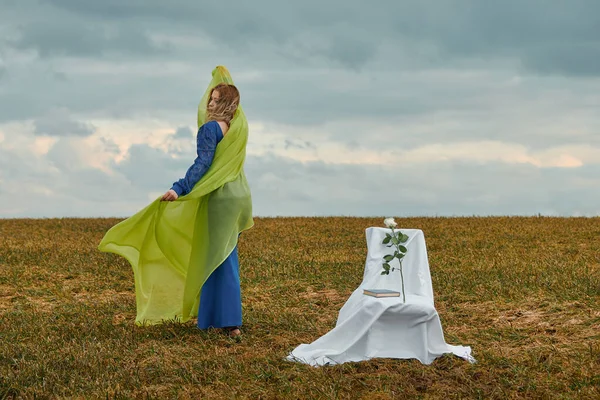 Sahada mavi elbiseli ve sarı peçeli güzel bir kadın. Açıklıkta bir sandalye var.