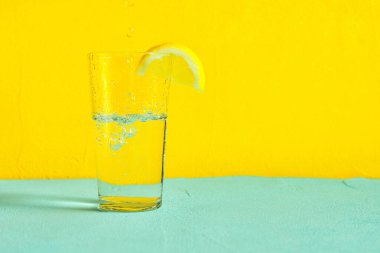 Sarı zemin üzerinde limon dilimleri olan bir bardak su.