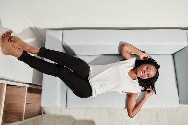 Afro-Amerikalı mutlu bir kız kanepeye uzanıp dinleniyor ve rüya görüyor. Üst Manzara. Genel giderler