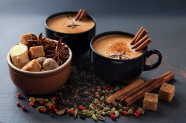 Masala çayı geleneksel sıcak Hint sütü siyah bardaklarda baharatlı içecek. Baharatlı tarçın çubukları, kakule kapsülleri, yıldız anason, şeker, hindistan cevizi, kişniş ve biber tohumları kahverengi seramik bardakta siyah taş tabakta. 