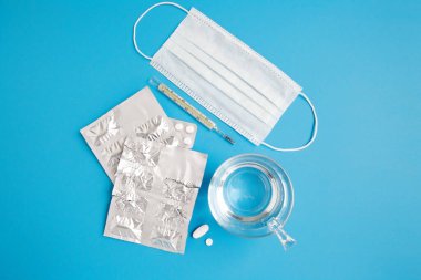 Bir bardak su, su toplayan haplar, maske ve mavi arka planda termometre.. 