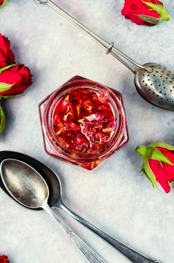 A brown ceramic tea kettle with a cup, rosebuds, and a glass of dried rose petals on a light background.