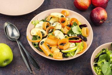 Çiğ elma, yapraklı yeşillikler ve tohumlarla süslenmiş havuç kurdeleleri içeren çiğ sebze salatası..