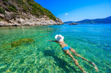 Afionas 'taki Porto Timoni plajında güneşlenen ve yüzen güneş şapkalı kız, Korfu, İyon adası, Yunanistan ve Avrupa' da kristal berraklığında masmavi suyu olan bir cennet.