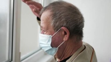 Koruyucu maskeli yaşlı adam pencereden dışarı bakar ve elini sallar, kendini izole eden 4K