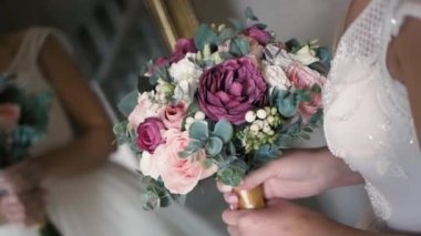 Beyaz elbiseli bir gelin elinde bir buketle aynanın önünde duruyor, bir buketin yakın çekimi, gündüz, kapalı bir yerde.