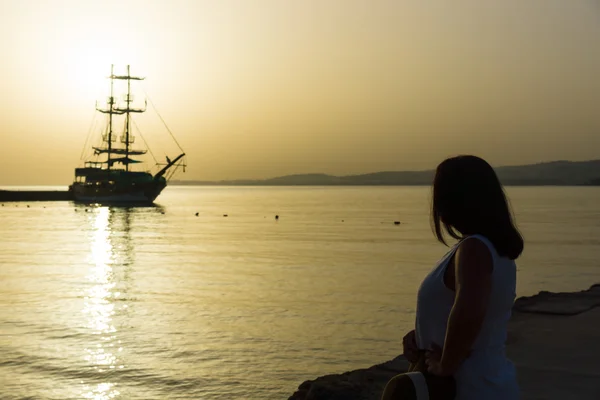 Belo pôr-do-sol sobre o mar. Barco à vela na baía. Mulher de pé na praia e olhando para o mar . — Fotografia de Stock