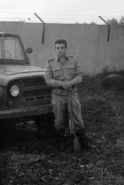 A soldier with a gun standing near army SUV UAZ-469. Film scan. Large grain — Stock Photo, Image