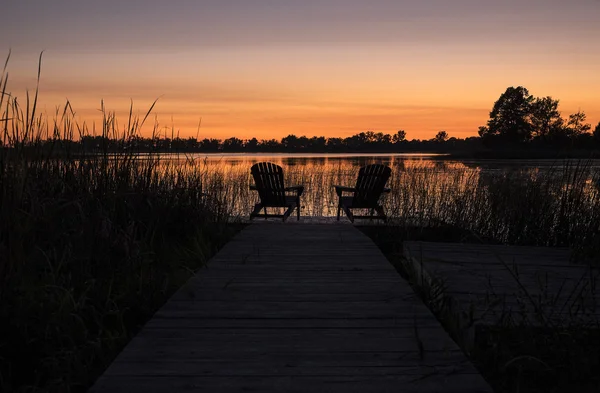 Silhueta de duas cadeiras Adirondack junto ao lago ao pôr do sol — Fotografia de Stock