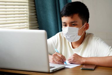 Asian boy wearing face mask and looked at laptop computer to study online, Online education via the internet during the COVID 19 epidemic, stay at home, quarantine concept. clipart