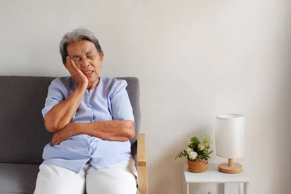 Elderly Asian woman with grey hair sits toothache on the sofa, Aging society and Various illnesses of the elderly and good health concept, with copy space for text.