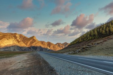 Scenic low angle view of mountain ridge. Highway in the foreground leading all the way up to the mountains. Beautiful cloudy sunset sky as a backdrop. Golden hour. Altai mountains, Siberia, Russia. clipart
