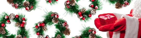 クリスマスの隣に紫色のリボンが付いた赤いギフトボックスを持っているサンタの手のショットを閉じる高い角度は赤い果実で花輪を松 クリスマスのギフトの概念 バナーだ 白地に隔離された — ストック写真