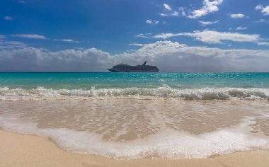 Half Moon Cay, Bahamas - February 19, 2020: Carnival Freedom anchoring by Half Moon Cay island. Gorgeous turquoise water splashing in the foreground. Cloudy blue sky in the background.