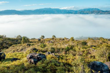 Geniş bir dağ arazisi, aşağıdaki vadiyi kaplayan dalgalı bir beyaz bulut denizini, berrak mavi gökyüzünün altında yükselen engebeli tepeleri, bir durgunluk ve yükseklik hissi uyandırıyor.