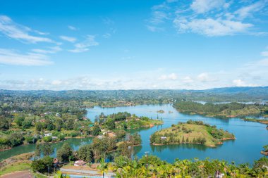 Yeşil adaları ve yarımadaları olan Guatap rezervuarları berrak mavi sulara gömülmüş. Parlak bir gökyüzünün altındaki ormanlık tepelere doğru uzanan bulutlar sakin ve canlı.