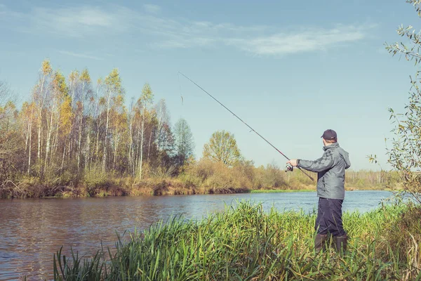 男は釣り竿を持って岸に立っている 釣り竿を持った上着を着た釣り人が川岸に立っている — ストック写真