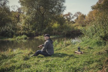 Adam ateşin yanındaki nehir kıyısında bir fincan kahveyle tek başına oturur. Doğada dinlen. Güneşli bir günde, bir kamp ateşinin yanında tek başına oturan bir adam.