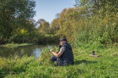 Elinde kupa olan genç adam nehrin kenarında oturuyor. Açık havada yürüyüş..