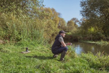 Elinde kupa olan genç adam nehrin kenarında oturuyor. Açık havada yürüyüş..