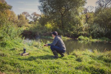 Adam çimlerin üzerinde oturur ve ateşin üzerinde kahve yapar. Nehir kenarındaki turizm. Erkek turist ateşte kahve yapıyor..