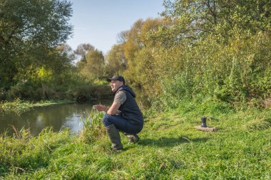 Elinde kupa olan genç adam nehrin kenarında oturuyor. Açık havada yürüyüş..