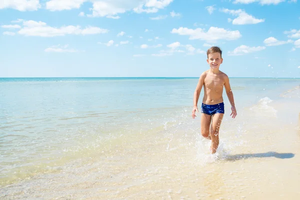 Netter kleiner Junge, der am Strand durchs Wasser rennt — Stockfoto