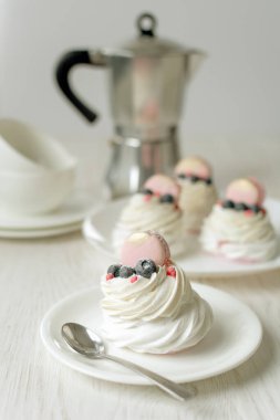 Hayat boyu Pavlova kremalı pasta, beyaz arka planda süslemeli makaron. Çay fincanı ve kahve makinesi ahşap masada.
