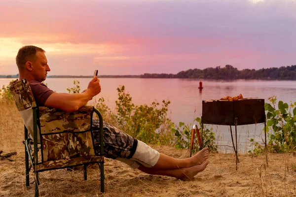 El hombre con teléfono celular en la mano está disfrutando de un paisaje increíble, mientras está sentado en la playa y asando una barbacoa — Foto de Stock