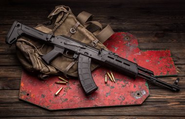 Modern bir vücut kitinde klasik bir Sovyet tüfeği AK-47. Karabina ve fişekler askeri sırt çantasının yanında ve hedef vurulmak üzere. Silahlar ahşap bir masada..